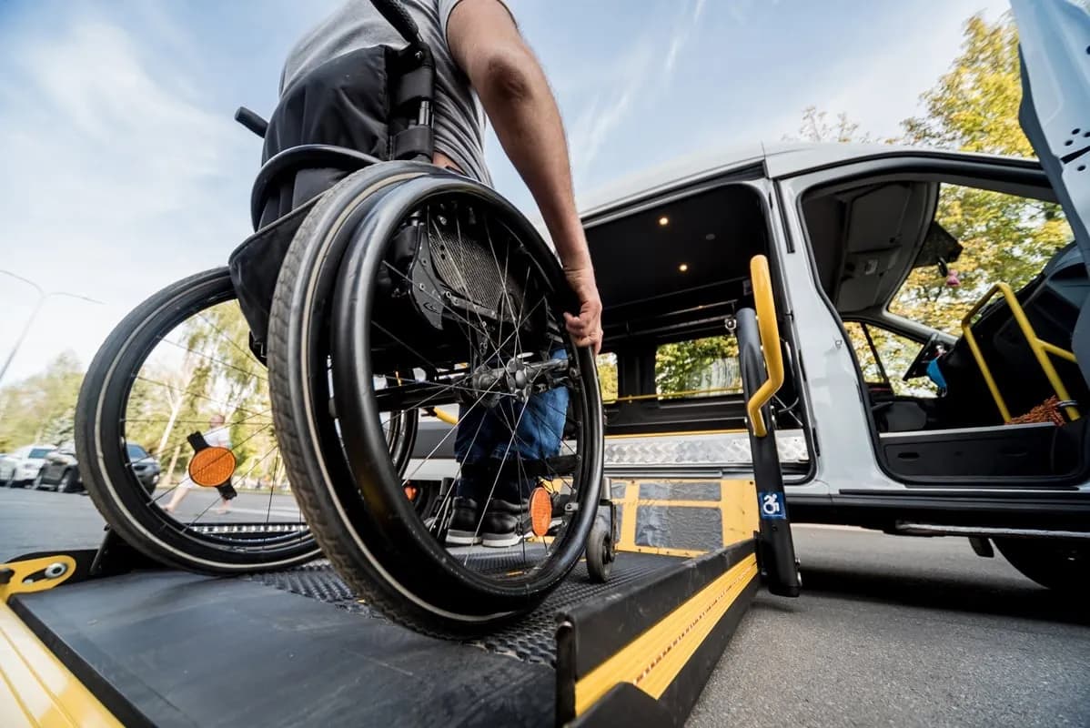 A person in a wheelchair is shown from the back, preparing to board an accessible van via a ramp with a yellow safety stripe.